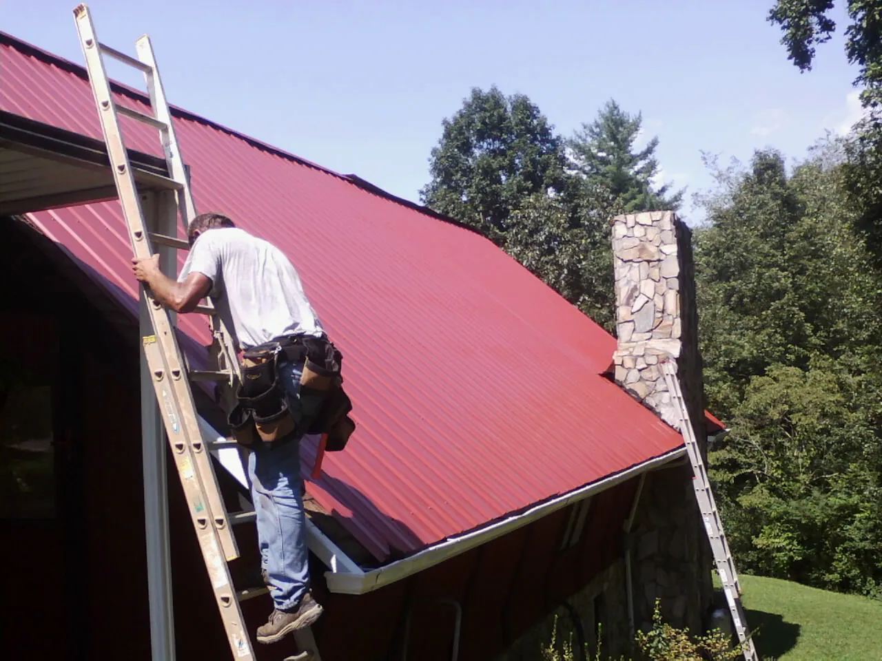 Roofer on a ladder beside a red metal roof and stone chimney — Soffit & Fascia Repair by Roofing Brothers in Colusa